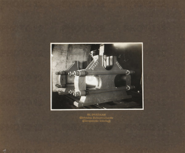 Sf. Pandaan Cast Steel Mill Stands (European delivery), album page, photograph, Endpaper with a photo of a mill stand. An employee is standing behind the machine. Manufactured or imported by Machinefabriek Braat and intended for the Pandaan Sugar Factory. Part of the photo album Machinefabriek Braat Soerabaia-Djocja-Tegal, ca. 1916-1924., Surabaya, 1916 - 1924, photographer, Java, East Java, paper, pasteboard, gelatin silver print, developed gelatin silver print, height 103 mm x width 143 mm, height 240 mm x width 290 mm, Person, NV Machinefabriek Braat, Suikerfabriek Pandaan, colonial system, Asiatic races and peoples (INDONESIANS), machines, parts of machines, tools and appliances, economic history, overseas history, sugar industry, company history, Pandaan, Period, A black-and-white photograph mounted on a dark textured background shows a large industrial metal component with curved and rectangular shapes, secured by long bolts and flanged clamps, resting on wooden pallets; a man is...