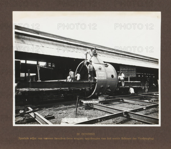 Sf. Gending, Special mode of transport suspended between two wagons of the first body of the Evaporation, album page, photograph, Album page with a photograph of the preparation for rail transport of an evaporation body. Produced by Machinefabriek Braat in Surabaya and intended for the Gending Sugar Factory. Part of the photo album Machinefabriek Braat Soerabaia-Djocja-Tegal, ca. 1916-1924., Surabaya, 1916 - 1924, photographer, Java, East Java, paper, pasteboard, gelatin silver print, developed gelatin silver print, height 162 mm x width 210 mm, height 240 mm x width 290 mm, Person, NV Machinefabriek Braat, Gending Sugar Factory, colonial system, machines, parts of machines, tools and appliances, freight train, economic history, overseas history, sugar industry, company history, Period, A black and white photograph showing a large cylindrical metal boiler-like structure mounted on a flat railcar platform with men working around and on it; the structure has riveted seams and circular op...
