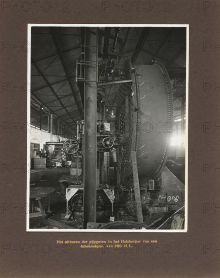 Drilling out pipe holes in the heating element of a 200 HL tubular cooking pot, album page, photograph, Album page with a photo of the operation of a machine. Produced by Machinefabriek Braat. Part of the photo album Machinefabriek Braat Soerabaia-Djocja-Tegal, ca. 1916-1924., Surabaya, 1916 - 1924, photographer, Java, East Java, paper, pasteboard, gelatin silver print, developing gelatin silver print, height 210 mm x width 162 mm, height 240 mm x width 290 mm, Person, NV Machinefabriek Braat, colonial system, factory-interior, machine shop, freight train, economic history, overseas history, sugar industry, company history, Period, A black and white photograph shows the interior of a large industrial workshop with a high pitched roof and visible metal trusses, a tall vertical metal column in the foreground with attached platforms and ladders, and a large circular metal drum with a riveted rim positioned to the right; various mechanical components, gears, pulleys, and handwheels are mou...