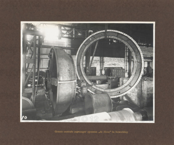 Large central juice collector system 'de Haan' in operation, album page, photograph, Album page with a photo of a machine part. Manufactured by Machinefabriek Braat. Part of the photo album Machinefabriek Braat Soerabaia-Djocja-Tegal, ca. 1916-1924., Surabaya, 1916 - 1924, photographer, Java, East Java, paper, pasteboard, gelatin silver print, developing gelatin silver print, height 157 mm x width 207 mm, height 240 mm x width 290 mm, Person, NV Machinefabriek Braat, colonial system, factory-interior, machine shop, machines, parts of machines, tools and appliances, economic history, overseas history, sugar industry, company history, Period, A black and white photograph shows an industrial workshop interior with large metal machinery and components. A large circular metal ring with bolt holes dominates the right side, suspended from a chain attached above. To the left of the ring is a curved metal housing and visible gears and shafts. Various cylindrical metal parts and pipes lie on the...