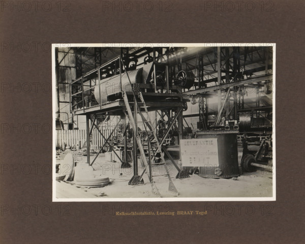 Lime milk installation. Delivery Braat Tegal, album page, photograph, Album page with a photo of an installation. Manufactured by Machinefabriek Braat. Part of the photo album Machinefabriek Braat Soerabaia-Djocja-Tegal, ca. 1916-1924., Tegal (city), 1916 - 1924, photographer, Java, Central Java, paper, pasteboard, gelatin silver print, developed gelatin silver print, height 146 mm x width 207 mm, height 240 mm x width 290 mm, Person, NV Machinefabriek Braat, colonial system, factory-interior, machine shop, machines, parts of machines, tools and appliances, economic history, overseas history, sugar industry, company history, Period, A black and white photograph shows the interior of an industrial workshop with a large elevated metal structure supported by angled steel legs and cross bracing; the structure includes a riveted cylindrical drum encased in a rectangular frame with a ladder leading up to the platform. Nearby are assorted machine components including large wheels, pulleys, ge...