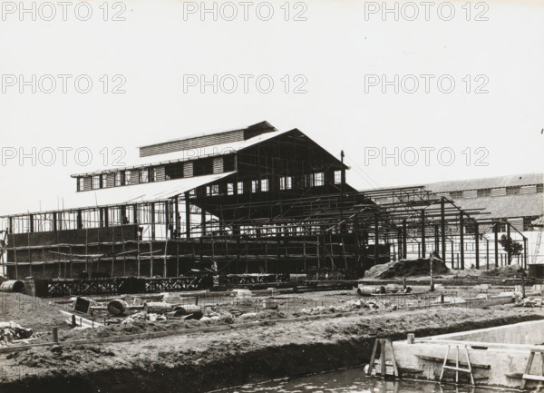 Marine Establishment, Surabaya, Submarine Repair Workshop, album page, photograph, Album page with a photo of the skeleton of a Navy submarine workshop in Surabaya, under construction. Part of the photo album Machinefabriek Braat Soerabaia-Djocja-Tegal, ca. 1916-1924., 1916 - 1924, photographer, Java, East Java, paper, pasteboard, gelatin silver print, developed gelatin silver print, height 179 mm x width 230 mm, height 240 mm x width 290 mm, Person, NV Machinefabriek Braat, Royal Netherlands Navy, navy, making the carcass (, building activities), colonial system, submarine, economic history, overseas history, architecture, business history, Period, A black-and-white photograph shows a large industrial building under construction with exposed steel framework and scaffolding, a multi-tiered roof structure with windows and open sections, and temporary supports and beams surrounding the structure; in the foreground there is a muddy construction site with scattered barrels, wooden planks, ...