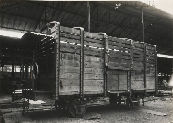 State Railways on Java, High open goods wagon type H. of which 230 units were delivered, album page, photograph, Album page with a photo of a type of railway wagon. Manufactured by Machinefabriek Braat and delivered to the State Railways on Java. Part of the photo album Machinefabriek Braat Soerabaia-Djocja-Tegal, ca. 1916-1924., Surabaya, 1916 - 1924, photographer, Java, East Java, paper, pasteboard, gelatin silver print, developed gelatin silver print, height 155 mm x width 205 mm, height 240 mm x width 290 mm, Person, Maatschappij tot Exploitatie van Staatsspoorwegen, NV Machinefabriek Braat, colonial system, railway wagon, factory-interior, machine shop, freight train, economic history, overseas history, sugar industry, company history, Period, A black and white photograph shows a wooden freight wagon with vertical and horizontal planks, metal reinforcements, hinges, and latches, positioned on railway wheels and tracks inside a large covered industrial shed with a corrugated roof a...