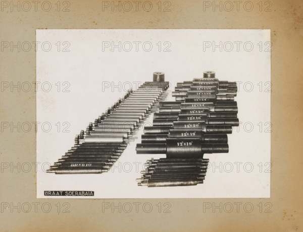 Exhibition of machine parts, album page, photograph, Album page with a photo of an exhibition of machine parts. Part of a photo album with photos of machines and constructions of the NV Machinefabriek Braat in Soerabaja, Soekaboemi and Tegal from the period 1931-1937., Surabaya, 1931 - 1937, photographer, Java, East Java, paper, pasteboard, gelatin silver print, developing gelatin silver print, height 174 mm x width 240 mm, height 250 mm x width 320 mm, Person, NV Machinefabriek Braat, parts of machines, economic history, overseas history, company history, Period, A rectangular white sheet is centered on a textured beige background, showing a large array of cylindrical metal rods and rollers arranged in parallel diagonal rows across the sheet, with one group of darker, thicker cylinders on the right and a group of thinner, lighter cylinders on the left; the cylinders vary in length and diameter and some have small protruding ends or textured surfaces, with two larger cylindrical pieces...