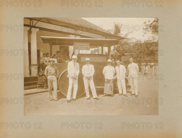 Roller transport, album page, photograph, Album page with a photo of a roller transported from Surabaya to Tasikmalaya in West Java. Dutch and Indonesian men are posing by the roller; this is likely in Tasikmalaya. Part of a photo album with photos of machines and constructions of the NV Machinefabriek Braat in Surabaya, Sukabumi, and Tegal from the period 1931-1937., Java, 1931 - 1937, photographer, paper, pasteboard, daylight gelatin silver print, height 167 mm x width 232 mm, height 250 mm x width 320 mm, Person, NV Machinefabriek Braat, other motor-vehicles not meant or mentioned above, economic history, overseas history, business history, Period, A sepia photograph showing a group of people standing in front of a large vehicle with a visible wheel and a canopy, with a building featuring columns and a tiled roof behind them; several men in light-colored suits and hats stand in a row at the front, alongside a man in a darker work shirt and a man wearing a patterned skirt and light j...