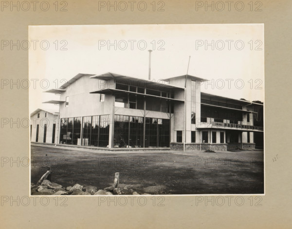 Pasir-Malang Industrial Building, album page, photograph, Album page with a photo of the industrial building of the tea and cinchona company Pasir-Malang, near Pangalengan, circa 1936. Part of a photo album with photos of machines and constructions of the NV Machinefabriek Braat in Surabaya, Sukabumi, and Tegal from the period 1931-1937., West Java, 1936, photographer, Java, paper, pasteboard, gelatin silver print, height 169 mm x width 233 mm, height 250 mm x width 320 mm, Person, NV Machinefabriek Braat, factory-building, economic history, overseas history, tea culture, company history, cinchona culture, Pangalengan, Period, 1931 - 1937, A grayscale photograph of a multi-story industrial building with large vertical glass windows on the ground level and smooth concrete panels above, featuring cantilevered roof overhangs and exposed structural supports; a tall narrow tower with vertical windows rises near the center-right and a thin smokestack extends upward behind the roofline; the f...