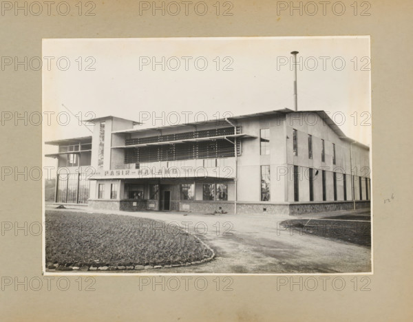 Pasir-Malang Tea and Cinchona Enterprise, album page, photograph, Album page with a photo of the front of a building of the Pasir Malang tea and cinchona enterprise near Pengalengan, built in 1936. Part of a photo album with photos of machines and constructions of the NV Machinefabriek Braat in Surabaya, Sukabumi, and Tegal from the period 1931-1937., West Java, 1936, photographer, Java, paper, pasteboard, gelatin silver print, height 167 mm x width 233 mm, height 250 mm x width 320 mm, Person, NV Machinefabriek Braat, factory-building, economic history, overseas history, tea culture, company history, cinchona culture, Period, 1931 - 1937, A black and white photograph of a two-story building with a flat overhanging roof and a tall cylindrical chimney rising above one corner, the facade featuring large vertical windows and a ground-level entrance recessed beneath a canopy, a horizontal band of louvered or slatted panels across the upper level, a tower-like projection on one side with na...