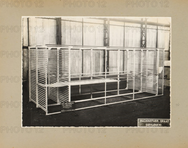 Paint lamination racks, album page, photograph, Album page with a photo of 'fixed paint lamination racks with rollable Hessian Cloth', November 19, 1935. Part of a photo album with photos of machines and constructions of the NV Machinefabriek Braat in Surabaya, Sukabumi and Tegal from the period 1931-1937., Sukabumi, 1935-11-19, photographer, Java, West-Java, paper, pasteboard, gelatin silver print, developing gelatin silver print, height 168 mm x width 227 mm, height 250 mm x width 320 mm, Person, NV Machinefabriek Braat, factory-interior, machine shop, machines, parts of machines, tools and appliances, economic history, overseas history, company history, Period, 1931 - 1937, A black and white photo shows a large metal shelving structure with multiple horizontal slats and rectangular frames arranged in rows, placed on a dark floor inside a corrugated metal-walled interior with vertical support beams and truss-like columns; the shelving has open grid sides and stacked tray-like levels,...