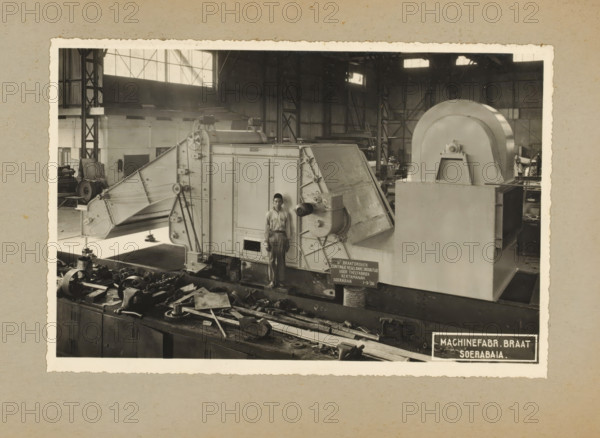 Braat dryer for the Kertamanah tea factory, album page, photograph, Album page with a photo of a drying machine, manufactured in 1936 and intended for the Kertamanah tea factory. Part of a photo album with photos of machines and constructions of the NV Machinefabriek Braat in Surabaya, Sukabumi and Tegal from the period 1931-1937., Surabaya, 1936-09-01, photographer, Java, East Java, paper, pasteboard, gelatin silver print, developing gelatin silver print, height 153 mm x width 230 mm, height 250 mm x width 320 mm, Person, NV Machinefabriek Braat, machines, parts of machines, tools and appliances, economic history, overseas history, tea culture, company history, Period, 1931 - 1937, A black and white photograph shows an industrial workshop interior with a large metal machine occupying the center and right side, featuring a rectangular body, angled panels, exposed gears, pulleys, and a large curved housing on the right; a man in work clothes and a cap stands in front of the machine agai...