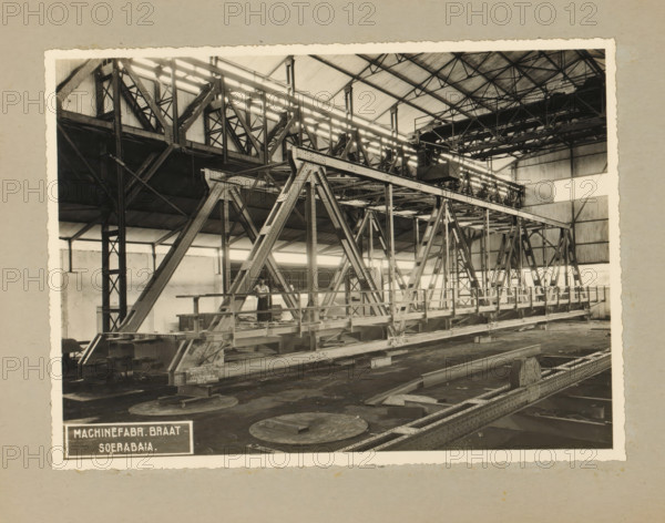 Railway bridge, album page, photograph, Album page with a photo of a truss bridge (railway bridge with footbridge) intended for the State Railways, January 29, 1937. Part of a photo album with photos of machines and constructions of the NV Machinefabriek Braat in Surabaya, Sukabumi, and Tegal from the period 1931-1937., Surabaya, 1937-01-29, photographer, Java, East Java, paper, pasteboard, gelatin silver print, developing gelatin silver print, height 168 mm x width 228 mm, height 250 mm x width 320 mm, Person, Company for the Exploitation of State Railways, NV Machinefabriek Braat, railway, train, factory-interior, machine shop, economic history, overseas history, bridge construction, company history, Period, 1931 - 1937, A sepia-toned photograph of an industrial interior showing a large metal truss bridge structure supported by triangular steel frames and riveted beams inside a high-roofed building with exposed roof trusses and corrugated wall panels; walkways and railings run along ...