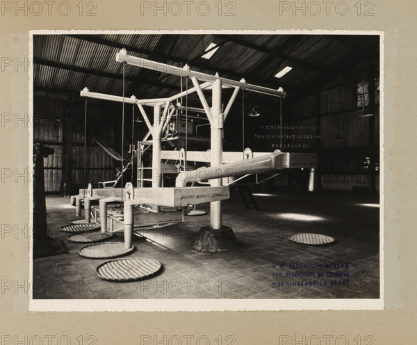 Installation, album page, photograph, Album page with a photo of an installation with conveyor belts. Designed for or by NV Technisch Bureau Van Assendelft De Coningh. Part of a photo album with photos of machines and structures of the NV Machinefabriek Braat in Surabaya, Sukabumi, and Tegal from the period 1931-1937., name: 'NV Technisch Bureau Van Assendelft De Coningh Sukabumi', name: 'NV Technisch Bureau Van Assendelft De Coningh Machinefabriek Braat', 1931 - 1937, photographer, Java, West-Java, paper, pasteboard, gelatin silver print, height 170 mm x width 216 mm, height 250 mm x width 320 mm, Person, NV Machinefabriek Braat, factory-interior, machine shop, machines, parts of machines, tools and appliances, economic history, overseas history, company history, Period, A black and white photograph of an industrial interior showing a large metal framework with horizontal beams and vertical supports, pipes and rods connected to a central column, suspended lights hanging from the ceili...