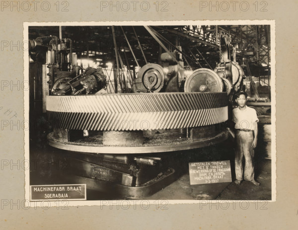 Gear, album page, photograph, Album page with a photo of a cast steel gear being worked on the milling machine in 1937 and intended for the Gempolkerep Sugar Factory. Part of a photo album with photos of machines and constructions of the NV Machinefabriek Braat in Surabaya, Sukabumi, and Tegal from the period 1931-1937., Surabaya, 1937-03-01, photographer, Java, East Java, paper, pasteboard, gelatin silver print, developing gelatin silver print, height 168 mm x width 227 mm, height 250 mm x width 320 mm, Person, NV Machinefabriek Braat, parts of machines, economic history, overseas history, sugar industry, company history, Period, 1931 - 1937, A black-and-white photograph showing a large industrial gear with diagonal toothed ridges mounted on a circular base, surrounded by machinery components including pulleys, chains, gears, and structural beams; a man wearing a light-colored short-sleeved shirt and trousers stands beside the gear near the right edge of the image; the background reve...