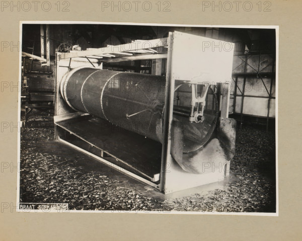 Drum, album page, photograph, Album page with a photo of a drum for a machine. Part of a photo album with photos of machines and constructions of the NV Machinefabriek Braat in Surabaya, Sukabumi, and Tegal from the period 1931-1937., 1931 - 1937, photographer, Java, West-Java, paper, pasteboard, gelatin silver print, developing gelatin silver print, height 169 mm x width 232 mm, height 250 mm x width 320 mm, Person, NV Machinefabriek Braat, factory-interior, machine shop, machines, parts of machines, tools and appliances, economic history, overseas history, company history, Period, A black and white photograph of an industrial interior showing a large cylindrical metal drum with a mesh surface mounted horizontally within a rectangular metal frame, supported by vertical posts and brackets, with a curved metal chute attached to one end of the drum and a clamping mechanism visible nearby; the drum sits above a flat tray or conveyor and leaves scattered across the floor; background elemen...
