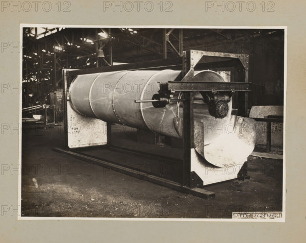 Drum, album page, photograph, Album page with a photo of a drum for a machine. Part of a photo album with photos of machines and constructions of the NV Machinefabriek Braat in Surabaya, Sukabumi, and Tegal from the period 1931-1937., 1931 - 1937, photographer, Java, West-Java, paper, pasteboard, gelatin silver print, developed gelatin silver print, height 172 mm x width 236 mm, height 250 mm x width 320 mm, Person, NV Machinefabriek Braat, factory-interior, machine shop, machines, parts of machines, tools and appliances, economic history, overseas history, company history, Period, A large cylindrical metal drum with a textured surface is mounted horizontally within a rectangular metal frame, visible from an angled front view inside a spacious industrial building. The drum shows segmented reinforcement bands and is supported by padded cradle blocks at each end, with a central shaft and bearing assembly projecting through the frame. The frame includes vertical and horizontal beams with ...