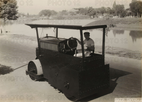 Roller with operator, album page, photograph, Album page with a photo of a roller with the Javanese operator. Part of a photo album with photos of machines and constructions of the NV Machinefabriek Braat in Surabaya, Sukabumi, and Tegal from the period 1931-1937., Surabaya, 1931 - 1937, photographer, Java, East Java, paper, pasteboard, gelatin silver print, height 166 mm x width 229 mm, height 250 mm x width 320 mm, Person, NV Machinefabriek Braat, machine driven by steam, other motor-vehicles not meant or mentioned above, economic history, overseas history, business history, Period, 1931 - 1938, A vintage road roller with a flat canopy and riveted metal panels is parked on a paved surface next to a body of water, a man in a light shirt sits at the controls holding the steering wheel, trees and low vegetation line the far bank with reflections visible in the calm water, and a single leafy tree stands near the water’s edge on the left side of the scene.
