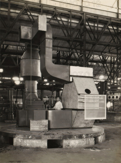 Fermentation machine, album page, photograph, A fermentation machine standing in a factory hall, September 27, 1934. Part of a photo album with photos of machines and constructions of the NV Machinefabriek Braat in Soerabaja, Soekaboemi, and Tegal from the period 1931-1937., Surabaya, 1934-09-11, photographer, Java, East Java, paper, pasteboard, gelatin silver print, developing gelatin silver print, height 240 mm x width 178 mm, height 250 mm x width 320 mm, Person, NV Machinefabriek Braat, factory-interior, machine shop, machines, parts of machines, tools and appliances, economic history, overseas history, company history, Period, 1931 - 1937, A large industrial interior with a tall metal structure composed of ducts and rectangular and curved panels rising from a rounded base, supported by a heavy circular concrete platform. The background shows a high corrugated metal roof with exposed steel trusses and beams creating a grid of diagonal and vertical lines. Bright overhead lights cast...