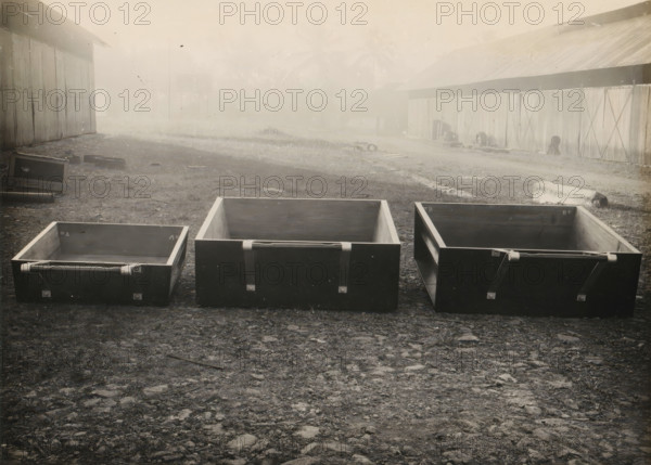 Three bins, album page, photograph, Three bins or drawers, parts of a machine. Part of a photo album with photos of machines and constructions of the NV Machinefabriek Braat in Surabaya, Sukabumi and Tegal from the period 1931-1937., Sukabumi, 1931 - 1937, photographer, Java, West-Java, paper, pasteboard, gelatin silver print, developed gelatin silver print, height 175 mm x width 234 mm, height 250 mm x width 320 mm, Person, NV Machinefabriek Braat, parts of machines, economic history, overseas history, company history, Period, Three rectangular open wooden boxes sit on a rough dirt and gravel ground in the foreground, each with metal straps and latches on their outer faces; behind them a grassy area leads to two long corrugated metal buildings flanking an open space, the left building showing a large overhanging roof and the right building featuring a series of vertical panels and a sloped roof; scattered debris and a few small objects lie on the ground between the buildings; the scen...