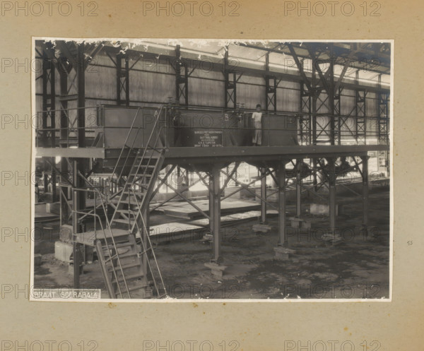 Platform with cooking pots, photograph, Album page with a photo of the interior of a factory hall containing a rack with cooking pots, ready for delivery to Guttabladbrei, October 30, 1934. Part of a photo album with photos of machines and constructions of the NV Machinefabriek Braat in Surabaya, Sukabumi, and Tegal from the period 1931-1937., Surabaya, 1934-10-30, photographer, Java, East Java, paper, pasteboard, gelatin silver print, developing gelatin silver print, height 166 mm x width 229 mm, height 250 mm x width 320 mm, Person, NV Machinefabriek Braat, factory-interior, machine shop, machines, parts of machines, tools and appliances, economic history, overseas history, company history, Period, 1931 - 1937, A sepia-toned photograph of an industrial interior showing a raised metal platform supported by a framework of girders and columns, with a set of metal stairs and handrails leading up to the platform and additional structural cross-bracing beneath; a person stands on the platf...