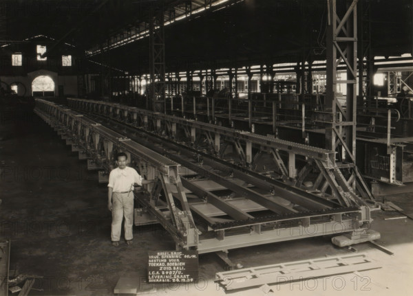 Cable bridge, album page, photograph, Album page with a photograph of a cable bridge manufactured in 1934 for the Dutch East Indies Central Procurement Service (ICA) and for installation in South Bali. Part of a photo album with photos of machines and constructions of the NV Machinefabriek Braat in Soerabaja, Soekaboemi, and Tegal from the period 1931-1937., Surabaya, 1934-12-24, photographer, Java, East Java, paper, pasteboard, gelatin silver print, developed gelatin silver print, height 174 mm x width 233 mm, height 250 mm x width 320 mm, Person, NV Machinefabriek Braat, factory-interior, machine shop, bridges by construction, economic history, overseas history, bridge construction, company history, Period, 1931 - 1937, A large indoor industrial space with high ceilings and structural beams, long metal framework assemblies arranged in parallel extending across the floor, a man standing in front of the nearest assembly wearing a light-colored shirt and trousers and resting one hand on...