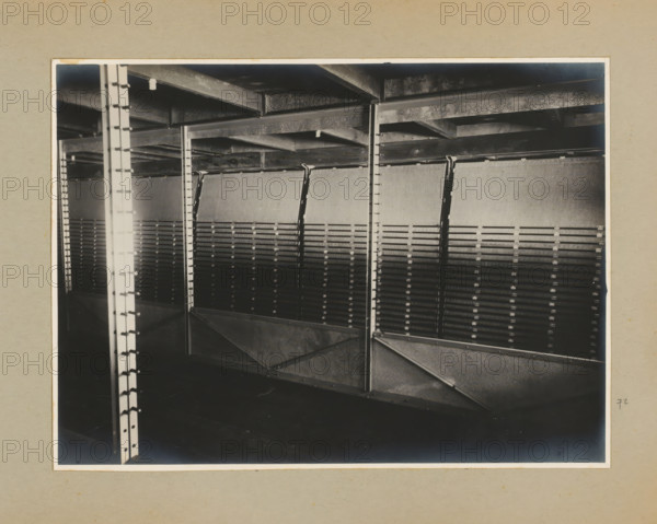 Installation, photograph, Album page with a photo of a machine or installation, inside a factory building. Part of a photo album with photos of machines and constructions of the NV Machinefabriek Braat in Surabaya, Sukabumi and Tegal from the period 1931-1937., Java, 1931 - 1937, photographer, paper, pasteboard, gelatin silver print, developed gelatin silver print, height 167 mm x width 227 mm, height 250 mm x width 320 mm, Person, NV Machinefabriek Braat, factory-interior, machine shop, economic history, overseas history, company history, Period, A black and white photograph framed by a light mat showing a metal interior structure with vertical support columns and horizontal beams, angled diagonal braces, and a series of evenly spaced slatted panels mounted between the supports, with visible rivets and bolts on the metal surfaces and a ceiling of exposed beams above, the lower area appearing dark and shadowed.
