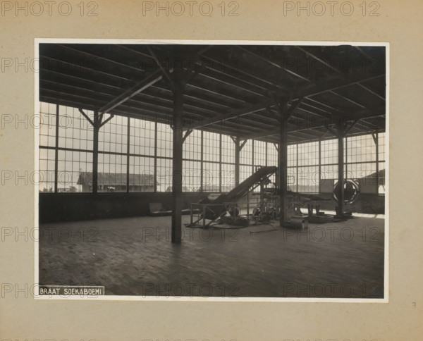 Factory hall with machine, album page, photograph, Album page with a photo of a factory building with a machine. Part of a photo album with photos of machines and constructions of the NV Machinefabriek Braat in Surabaya, Sukabumi and Tegal from the period 1931-1937., Sukabumi, 1931 - 1937, photographer, Java, West-Java, paper, pasteboard, gelatin silver print, developing gelatin silver print, height 173 mm x width 235 mm, height 250 mm x width 320 mm, Person, NV Machinefabriek Braat, factory-interior, machine shop, machines, parts of machines, tools and appliances, economic history, overseas history, company history, Period, A large interior space with high ceilings supported by dark vertical and diagonal wooden beams and a grid of horizontal ceiling joists, tall multi-pane windows lining the walls that let in light, a wooden floor with a worn parquet pattern, industrial machinery including a sloped conveyor-like apparatus and various metal components and supports clustered toward one ...