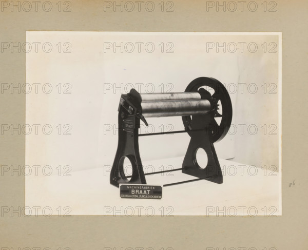 Roller press, album page, photograph, Album page with a photo of a type of roller press. Part of a photo album with photos of machines and constructions of the NV Machinefabriek Braat in Surabaya, Sukabumi, and Tegal from the period 1931-1937., Java, 1931 - 1937, photographer, paper, pasteboard, gelatin silver print, developing gelatin silver print, height 164 mm x width 230 mm, height 250 mm x width 320 mm, Person, NV Machinefabriek Braat, machines, parts of machines, tools and appliances, pressing (industrial process), economic history, overseas history, company history, Period, A photographic print of a mechanical device mounted on a plain light background, showing a pair of cylindrical metal rollers held between two dark metal A-shaped frames with circular cutouts at their bases, connected by horizontal bars; a large dark wheel with spokes is attached to one side of the rollers, and a lever-like handle is visible near the roller assembly; the image is bordered by a lighter rectangu...