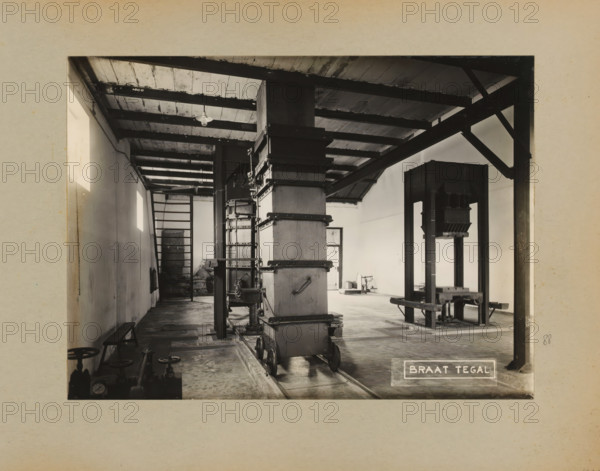 Factory hall, album page, photograph, Album page with a photo of the interior of a business space with chutes and presses. Part of a photo album with photos of machines and constructions of the NV Machinefabriek Braat in Surabaya, Sukabumi, and Tegal from the period 1931-1937., Tegal (city), 1931 - 1937, photographer, Java, Central Java, paper, pasteboard, gelatin silver print, gelatin silver developing print, height 169 mm x width 230 mm, height 250 mm x width 320 mm, Person, NV Machinefabriek Braat, factory-interior, machine shop, economic history, overseas history, company history, Period, A monochrome interior scene shows an industrial room with exposed wooden ceiling beams and a row of light fixtures, a tall vertical metal and wooden column mounted on small wheeled rails, adjacent machinery with metal frames and platforms, a ladder against the far wall, a bench along one side wall, various pipes and valves near the foreground floor, and a plain plastered wall with rectangular wind...