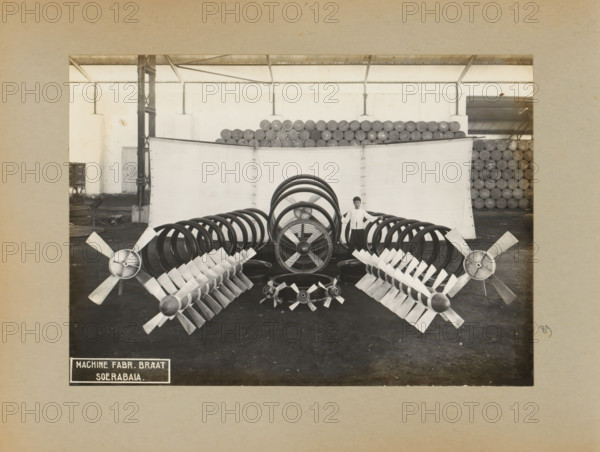 Fans, album page, photograph, Album page with a photo of a display of fans. Part of a photo album with photos of machines and constructions of the NV Machinefabriek Braat in Soerabaja, Soekaboemi and Tegal from the period 1931-1937., Surabaya, 1931 - 1937, photographer, Java, East Java, paper, pasteboard, gelatin silver print, developing gelatin silver print, height 165 mm x width 231 mm, height 250 mm x width 320 mm, Person, NV Machinefabriek Braat, machines, parts of machines, tools and appliances, ventilation systems, economic history, overseas history, company history, Period, A large indoor space with a high corrugated roof and exposed support columns contains stacks of cylindrical rolls arranged along the back wall, a tall light-colored partition in front of the stacks, and a central display of large circular metal rings and multiple propeller-like metal assemblies arranged symmetrically on the floor; a person stands behind the central rings, hands resting on two rings, and the f...