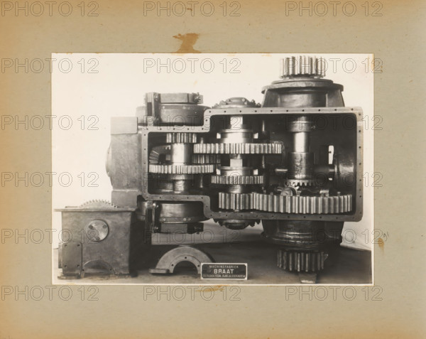 Machine, album page, photograph, Album page with a photo of a machine with gears. Part of a photo album with photos of machines and constructions of the NV Machinefabriek Braat in Surabaya, Sukabumi and Tegal from the period 1931-1937., Java, 1931 - 1937, photographer, paper, pasteboard, gelatin silver print, developed gelatin silver print, height 164 mm x width 228 mm, height 250 mm x width 320 mm, Person, NV Machinefabriek Braat, machines, parts of machines, tools and appliances, economic history, overseas history, company history, Period, A sepia-toned photograph mounted on a textured board shows a large industrial gearbox exposed to reveal its internal components, with multiple interlocking metal gears of various sizes fitted within a rectangular housing, cylindrical shafts, flanged bearing housings, and bolted joints, all rendered in polished and darkened metal tones; the housing has a thick rim with evenly spaced fastener holes and a rounded top section with a prominent gear atop...