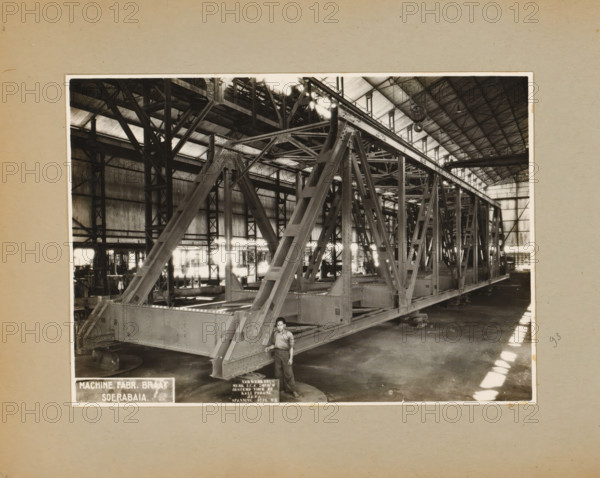 Truss bridge, album page, photograph, Album page with a photo of a factory hall with a truss bridge, intended to span the Kali Porong. Part of a photo album with photos of machines and structures of the NV Machinefabriek Braat in Surabaya, Sukabumi, and Tegal from the period 1931-1937., Surabaya, 1931 - 1937, photographer, Java, East Java, paper, pasteboard, gelatin silver print, developing gelatin silver print, height 160 mm x width 224 mm, height 250 mm x width 320 mm, Person, NV Machinefabriek Braat, bridge, factory-interior, machine shop, economic history, overseas history, bridge building, company history, Period, A sepia-toned photograph shows the interior of a large industrial building with a vaulted roof supported by a grid of metal trusses and beams; a massive riveted steel framework structure fills most of the central space, composed of angled supports, cross-bracing, and large horizontal sections resting on blocks, with visible rivet heads and flanged edges; the background r...