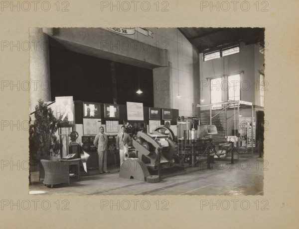 Men posing in the factory by a display of machines, album page, photograph, Album page with a photo of two European men posing in the factory by a display of machines, including those for the tea industry. On a wall, the city coat of arms of Bandung with the motto 'Ex Undis Sol'. Part of a photo album with photos of machines and structures of the NV Machinefabriek Braat in Surabaya, Sukabumi, and Tegal from the period 1931-1937., Bandung, 1931 - 1937, photographer, Java, West-Java, paper, pasteboard, gelatin silver print, developed gelatin silver print, height 160 mm x width 221 mm, height 250 mm x width 320 mm, Person, NV Machinefabriek Braat, director, boss (in modern industry), factory-interior, machine shop, machines, parts of machines, tools and appliances, economic history, overseas history, tea culture, company history, Period, A large indoor space with high walls and tall windows displaying light, rows of hanging pendant lights, and a corrugated roof; an exhibition area filled ...