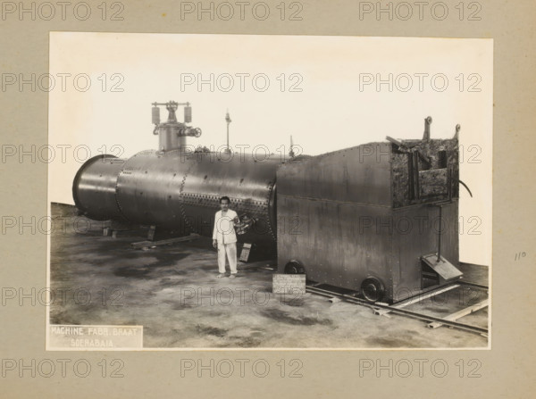 Locomobile boiler, album page, photograph, Album page with a photo of a locomobile boiler with superheater, manufactured December 23, 1935 and intended for Kertasarie. A man is posing by the boiler. Part of a photo album with photos of machines and constructions of the NV Machinefabriek Braat in Soerabaja, Soekaboemi, and Tegal from the period 1931-1937., Surabaya, 1935-12-23, photographer, Java, East Java, paper, pasteboard, gelatin silver print, developed gelatin silver print, height 160 mm x width 227 mm, height 250 mm x width 320 mm, Person, NV Machinefabriek Braat, machines, parts of machines, tools and appliances, economic history, overseas history, company history, Period, 1931 - 1937, A large riveted cylindrical metal boiler and an attached rectangular metal housing rest on wooden supports and rails inside a pale bordered frame, with a barefoot man in light-colored clothing standing in front of the cylindrical section holding a loose pipe fitting, the boiler surface showing sea...