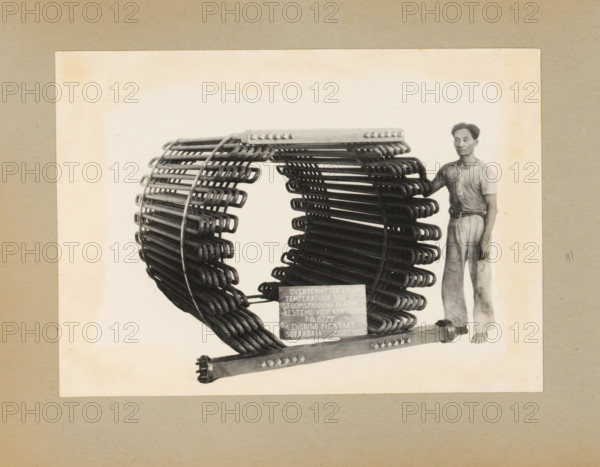 Superheater, album page, photograph, Album page with a photograph of a superheater, manufactured in 1935 and intended for (tea factory?) Kertasarie, December 23, 1935. A man poses standing next to the machine. Part of a photo album with photos of machines and constructions of the NV Machinefabriek Braat in Soerabaja, Soekaboemi, and Tegal from the period 1931-1937., Surabaya, 1935-12-23, photographer, Java, East Java, paper, pasteboard, gelatin silver print, developed gelatin silver print, height 161 mm x width 227 mm, height 250 mm x width 320 mm, Person, NV Machinefabriek Braat, machines, parts of machines, tools and appliances, economic history, overseas history, tea culture, company history, Period, 1931 - 1937, A sepia-toned photograph shows a large circular metal coil structure made of many curved rods arranged in concentric layers mounted on a horizontal base, with a man standing to the right of the structure wearing a short-sleeved shirt and loose trousers and resting one hand ...