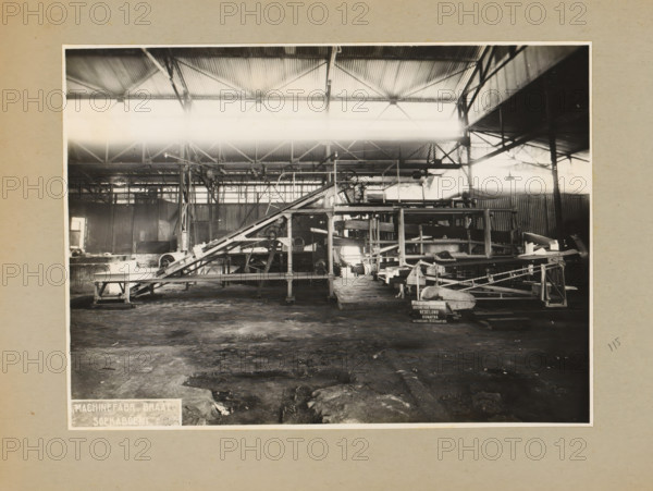 Tea sorting installation, album page, photograph, Album page with a photo of a tea sorting installation, manufactured in 1935 and intended for the Redelong company on Sumatra. Part of a photo album with photos of machines and structures of the NV Machinefabriek Braat in Surabaya, Sukabumi, and Tegal from the period 1931-1937., 1935-03-10, photographer, Java, West-Java, paper, pasteboard, gelatin silver print, developed gelatin silver print, height 166 mm x width 227 mm, height 250 mm x width 320 mm, Person, NV Machinefabriek Braat, factory-interior, machine shop, machines, parts of machines, tools and appliances, economic history, overseas history, tea culture, company history, Period, 1931 - 1937, A black and white photograph of an industrial interior showing a large open space with a corrugated metal roof supported by exposed beams and columns, a system of metal conveyors and elevated frameworks occupying the central area with angled chutes, rollers, and platforms, a dirt or rough fl...