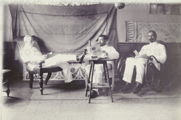 Group photo in the Gravenstraat, photograph, Group photo of Andries Boom, Dr. van Hall and Mr. van de Werk, in the house of Van Hall in the Gravenstraat in Paramaribo, October 1908. Part of the photo album of the Boom-Gonggrijp family in Suriname and Curaçao., inscription: 'Gravenstraat October 1908 P'bo', photographer: Andries Augustus Boom, Paramaribo (city), 1908-10, Andries Augustus Boom, paper, photography, height 113 mm x width 174 mm, Person, Constant Johann Jakob van Hall, overseas history, Period, 1908, Three men in light-colored clothing sit in chairs in a room with patterned fabric hung on the wall behind them, each holding a book or magazine. The man on the left reclines with his legs stretched toward the center, resting on a small table, and sits in an ornate wooden chair with a woven back. The man in the middle sits in a wicker chair, looking slightly to his left while holding an open magazine above a stack of papers on a small wooden table that has a glass bottle on its ...