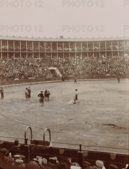 At the Bull Fight, Spain, photograph, Arena for bullfights in Valencia or Barcelona., 1905 - 1922, photographer, paper, gelatin silver print, developing gelatin silver print, height 105 mm x width 85 mm, Period, A large circular arena with a dirt floor fills the image, surrounded by tall tiered wooden stands packed with people; many spectators wear hats and there are railings and arches at the front of the stands. In the arena several figures stand near a bull, including individuals in traditional clothing and capes, while a few other people are scattered around the perimeter. The foreground shows the backs of seated spectators and the top of a barrier with metal rings and posts. The overall photograph has a sepia tone and a slightly faded, aged appearance.