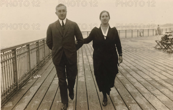 Photographs from the Estate of Isabel Wachenheimer, photograph, Eugen Wachenheimer (left) and his wife Else Wachenheimer-Moos (right) walking arm in arm on the quay of Scheveningen., date: 'Scheveningen August 1924.', Scheveningen, 1924-08, photographer, paper, gelatin silver print, developed gelatin silver print, height 90 mm x width 140 mm, Person, Eugen Wachenheimer, Else Wachenheimer-Moos, Period, A man and a woman walk side by side on a wooden boardwalk with a metal railing along the left edge; the man wears a dark three-piece suit with a white shirt and tie and has his hands behind his back, while the woman wears a dark dress or coat with a light collar and a necklace and has one arm linked through the man's arm and the other hanging by her side; the boardwalk shows parallel planks and worn patches, and in the background there are folding chairs and a distant figure near the railing with a hazy expanse beyond.