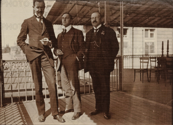 Eugen Wachenheimer as a young man with two unknown men outside under a canopy, 1906-1926, photograph, Eugen Wachenheimer as a young man with two unknown men outside under a canopy., 1906 - 1926, photographer, paper, gelatin silver print, developed gelatin silver print, height 90 mm x width 120 mm, Person, Eugen Wachenheimer, Period, Three men stand on a covered wooden balcony with a decorative metal railing. The men wear dark suits with waistcoats and ties; one man on the left leans with a hand in his pocket and the middle man holds something small between his fingers. The balcony has a slatted roof overhead and a tiled roof visible beyond the railing. In the background there are windows of a building and a couple of chairs and a small table near the railing. The photograph is sepia toned and shows fabric-like texture across the image.
