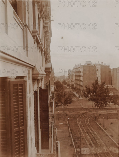 View from a balcony onto a street in an unknown city, 1905-1922, photograph, View from a balcony onto a street in an unknown city., 1905 - 1922, photographer, paper, gelatin silver print, developed gelatin silver print, height 125 mm x width 90 mm, Period, A sepia-toned photograph showing the facade of a multi-story building with shuttered windows and decorative stonework along a street lined with trees and tram tracks, the building's balconies and ornate cornices visible in the foreground while farther down the street other multi-story buildings with uniform windows and flat rooftops stand along the avenue, trees dot the sidewalk and a curved set of tracks runs toward the foreground, with lamp posts and a few indistinct figures near the distant buildings.