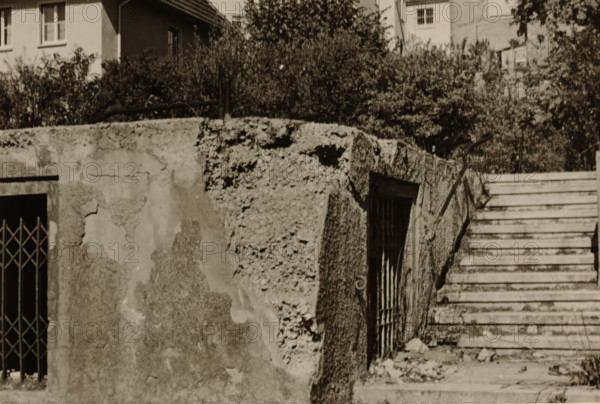 Part of a ruin with a sliding gate and a doorway in it, with a large house with a balcony in the background, with a tree and bushes around it, photograph, with a tree and bushes around it., 1945 - 1970, photographer, paper, gelatin silver print, developed gelatin silver print, height 75 mm x width 105 mm, Period, A sepia-toned scene showing a weathered concrete wall with areas of crumbling texture and peeling surface beside a set of worn stone steps leading upward; a metal barred gate set into a recessed opening is visible at the base of the wall and another barred window is set at an angle in the textured wall face; vegetation and dense shrubbery grow behind the wall, and building façades with windows and a tiled roof appear above the foliage in the background.