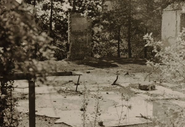 Part of a ruin, surrounded by dense vegetation of trees and shrubs, photograph, surrounded by dense vegetation of trees and shrubs., 1945 - 1970, photographer, paper, gelatin silver print, developed gelatin silver print, height 75 mm x width 105 mm, Period, A grayscale scene showing a partially cleared outdoor area with a broken brick chimney standing near the center, flanked by tall trees in the background and a tall rough wall fragment on the right edge. In the foreground there are scattered flat concrete slabs, small protruding metal rods and remnants of construction debris, with overgrown weeds and thin plant stems rising up. A blurred leafy bush appears on the left side and another blurred shrub on the right, partially obscuring the view, while shadows from the trees create dappled darker areas across the ground.