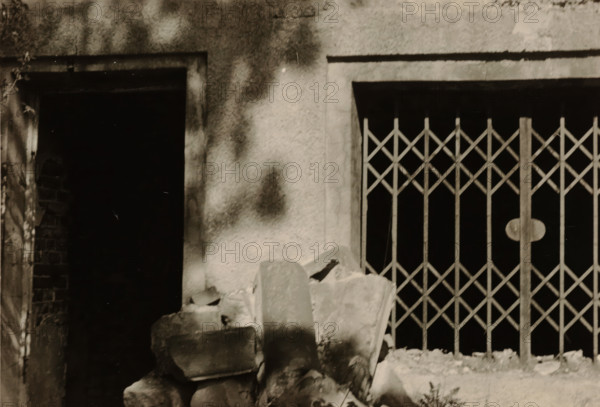 Enlargement of part of a ruin, with a sliding gate on the right and a doorway on the left, photograph, with a sliding gate on the right and a doorway on the left., 1945 - 1970, photographer, paper, gelatin silver print, developed gelatin silver print, height 75 mm x width 105 mm, Period, A cropped view of a textured wall with two rectangular openings, one dark open doorway on the left and a barred window with a diamond-pattern metal grille on the right, a pile of rough broken stones and rubble in front of the openings, a vertical column or post forming part of the grille with a round plate, and mottled light and shadow across the wall surface.