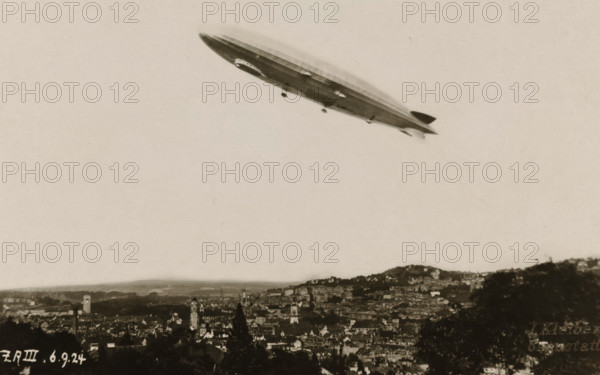 Flying Zeppelin over a city, photograph, Flying Zeppelin over a city., 1924-09-06, photographer, paper, gelatin silver print, developed gelatin silver print, height 85 mm x width 135 mm, Period, A large rigid airship floats above a sprawling cityscape, photographed from a distance; the airship has a streamlined elongated hull with a gondola underneath and small fins at the rear, shown against a pale sky. Below, the city fills the lower portion of the image with densely packed buildings, towers, and rooftops extending across rolling hills, with darker foreground trees framing the bottom edges. The overall image has a sepia tone and a slightly grainy texture.