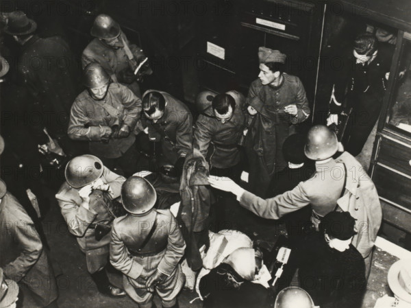 Dutch soldiers in London, photograph, Photo, taken from a certain height, with several (Dutch) soldiers stepping out of a train on a platform, with a piece of paper (carbon copy) with numbers, reverse side, with pen, explanation of those numbers: '1: Sergt. H. vd Togt, 2: Korp. J. v. Arkel, 3: HJ Brakema (Middelborst), 4: Sergt. DH Cevat, 5: Sergt. A. Domhof, 6: Private Edm. Eymael, 7: 2nd Lt. Mulders, 8: Sergt. H. Kahn, 9: 2nd Lt. Ebens, 10: Capt. L. vd Bergh, 11: Mrs. vd Bergh, 12: Mrs. Mulders., London (possibly), 1940 - 1944, photographer, London, materials by function, paper, ink, photography, writing (processes), height 14.7 cm x width 18.9 cm, Second World War, military history, England, Period, 1940 - 1945, A crowded black-and-white scene showing several uniformed men wearing helmets and coats gathered closely together, some facing inward and some turned toward the edges of the frame, with a few men bending over or reaching toward items on a surface; one man at right wears a di...