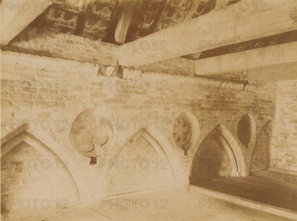 Arched windows and niches in the abbey church in Aduard, photograph, annotation: 'Aduard', annotation: 'Aduard, Arched windows and niches west of the NH church, and above the former school, June 1900', Aduard, 1900, photographer, Attributed to, paper, pasteboard, albumen print, height 173 mm x width 230 mm, parts of church interior: niche, A sepia-toned interior view of a brick structure showing a low wall with pointed arch openings and circular decorative stone elements above the arches, rough wooden beams crossing overhead, exposed rafters and roof tiles between the beams, uneven brickwork along the upper wall, and a wooden floor surface in the foreground.