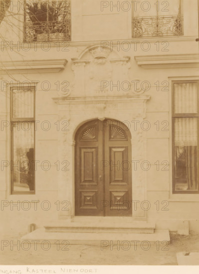 Portal of Nienoord Manor in Leek, photograph, annotation: 'Entrance of the castle (modern), Nienoord, in Leek province (Gr.), the small gate from 1678 has apparently been restored, April 1897', annotation: 'Entrance Nienoord Castle, Leek', Leek, 1897, photographer, Attributed to, paper, pasteboard, albumen print, height 228 mm x width 174 mm, gate, entrance, A double wooden door with carved rectangular panels and fan-shaped carvings in the upper sections sits within an arched stone doorway that has ornate carved details and a decorative crest above, flanked by tall windows with curtains and reflective glass, stone steps leading up to the entrance, smooth stone facade with horizontal joint lines, and parts of bare tree branches visible against the building.