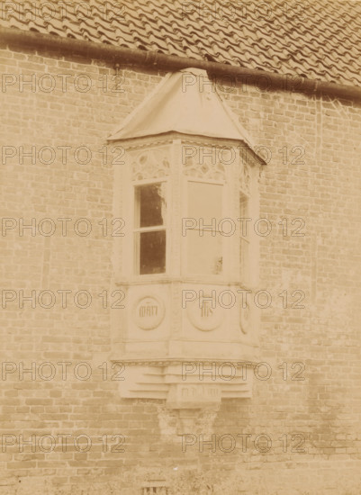 Bay window on the north facade of the Ter Apel Monastery, photograph, annotation: 'Ter Apel', annotation: 'Ter Apel, Bay window on the north facade of the monastery, Sept 1893', Ter Apel, 1893, photographer, Attributed to, paper, pasteboard, albumen print, height 232 mm x width 170 mm, bay-window, oriel, A sepia-toned photograph of a brick wall with a projecting bay window feature topped by a small pitched roof, the bay window has decorative carved panels above the glass panes and circular carved medallions below, a gutter and tiled roof run along the top of the image, and the brickwork shows varied tones and weathering.