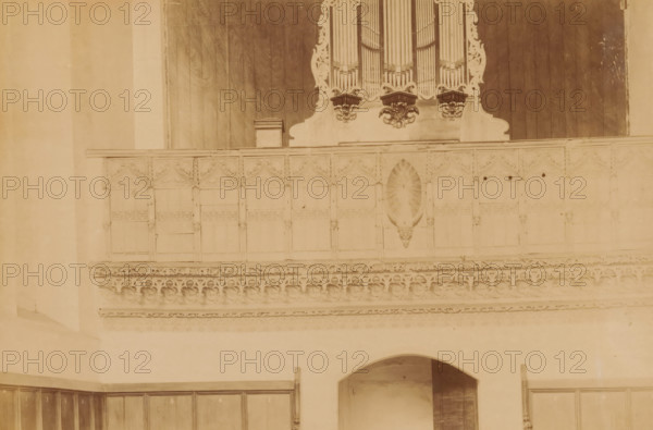 Choir screen in the Monastery Church Ter Apel, photograph, annotation: 'Ter Apel', annotation: 'Ter Apel, Jubilee in the church, total view, Sept 1893', Ter Apel, 1893, photographer, Attributed to, paper, pasteboard, albumen print, height 158 mm x width 235 mm, interior of church, rood-screen, choir-screen, iconostasis, A sepia-toned photograph showing an interior wall with decorative wooden paneling and ornate carved stonework; at the top center there is a section of an organ with visible vertical pipes and carved scrollwork beneath them, flanked by dark wooden panels. Below the organ a high balcony-like wooden railing features repeated carved panels with pointed arch motifs and a central carved niche with a sunburst or shell-shaped relief. Beneath the railing a horizontal band of intricate stone tracery and rounded quatrefoil-like patterns runs across the wall. Lower on the wall an arched opening leads into a recessed space, and wooden cupboard doors or panels line the lower portions...