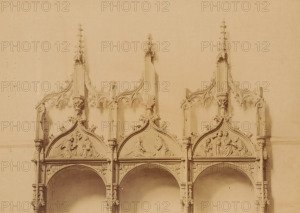 Details of the three stone seats in the Kloosterkerk Ter Apel, photograph, annotation: 'Ter Apel, Upper part of the 3 stone seats against the south wall of the choir of the church, Sept 1893', annotation: 'Ter Apel', Ter Apel, 1893, photographer, Attributed to, paper, pasteboard, albumen print, albumendruk, height 164 mm x width 231 mm, interior of church, sculpture (relief, sculpture), chair (stone), A carved stone architectural panel with three pointed ornamental gables sitting atop a series of arches, each gable containing shallow relief scenes of human figures in various poses; slender pinnacles rise above each gable with decorative crockets, tracery and small sculpted floral motifs along the vertical supports, and columns with capitals separating the arches below; the surface has a uniform warm sepia tone and shows slight variations in shading and some small speckled marks.