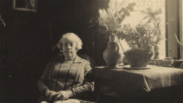 Portrait of Line van der Linden, photograph, photographer: circle of Cornelia Hendrika Jonker, Alkmaar, 1929, paper, gelatin silver print, developed gelatin silver print, height 63 mm x width 106 mm, Person, Line van der Linden, historical persons, interior of the house, An older woman with light-colored hair and round glasses sits in a chair, wearing a long-sleeved dress with vertical stripes and holding knitting needles and yarn in her hands, positioned next to a table covered with a cloth that holds a large ornate vase, a woven basket planter with tall foliage and flowers, and several small pots by a bright window.